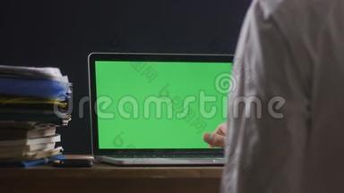 关闭人工作在笔记本电脑晚办公室与绿色屏幕。 在电脑前工作的商人
